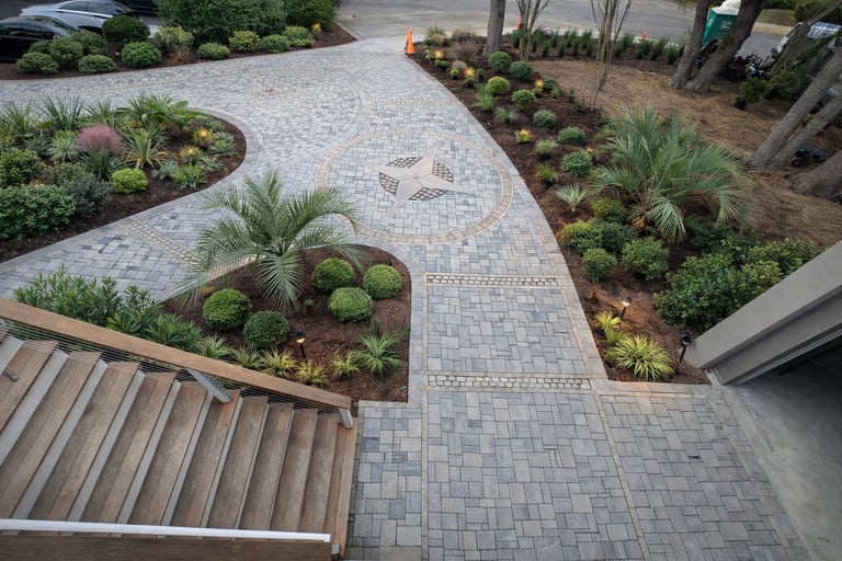 Aerial view of a modern landscaped patio with gray paver pathways, green plants and shrubs in mulched beds, and a metal roof visible in corner