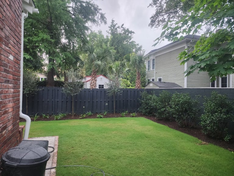 Backyard with lush green lawn, dark wooden fence, mature trees, brick house on left, and neighboring homes visible beyond fence