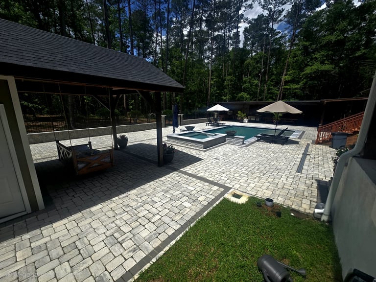 Modern backyard patio with pool, lounge chairs, pergolas, and pavers surrounded by trees
