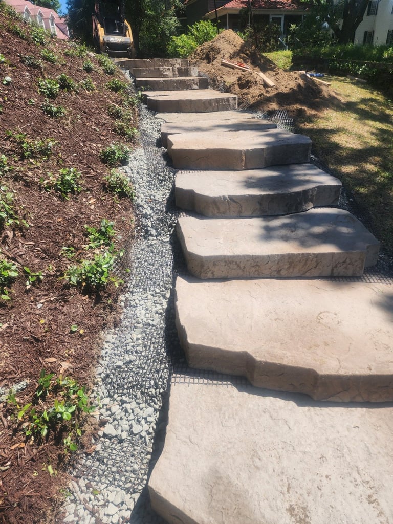 Stone stepping stones pathway descending a hillside with mulch and plants on left, surrounded by grass and trees
