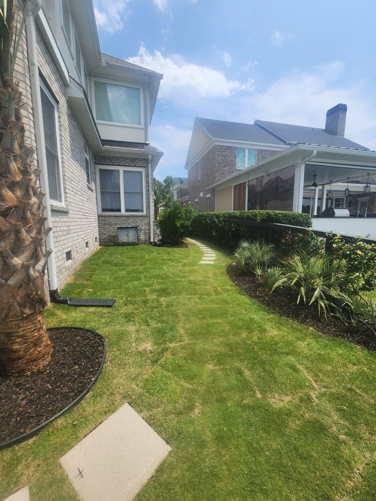 Well-maintained residential backyard with green lawn, brick houses, hedges, ornamental plants, and a pergola structure under clear sky