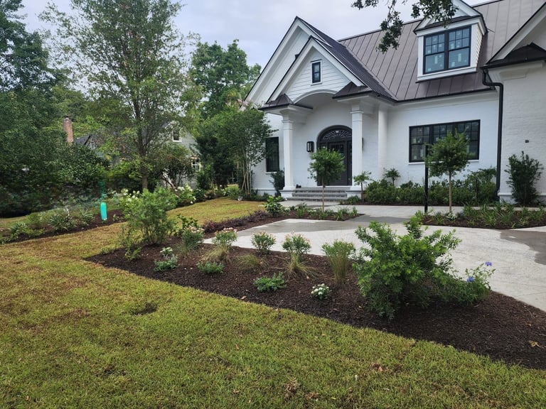 Modern white farmhouse with manicured front lawn, landscaped garden beds, and curved driveway surrounded by lush green trees